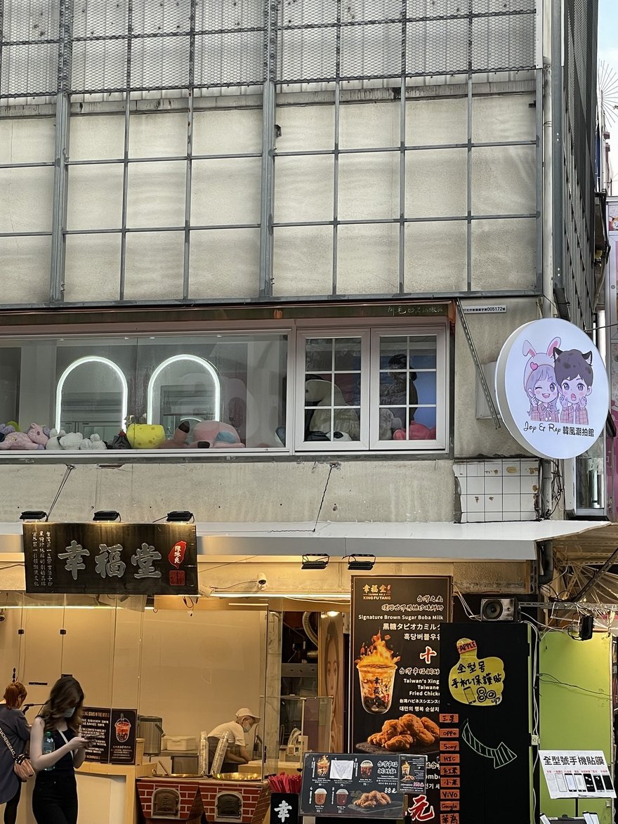 Joy&Ray 西門店的完整店面外觀，圓形燈箱招牌掛在騎樓外，二樓窗內是拱形燈鏡與娃娃堆，一樓是幸福堂黑糖珍奶與手機保護貼攤位