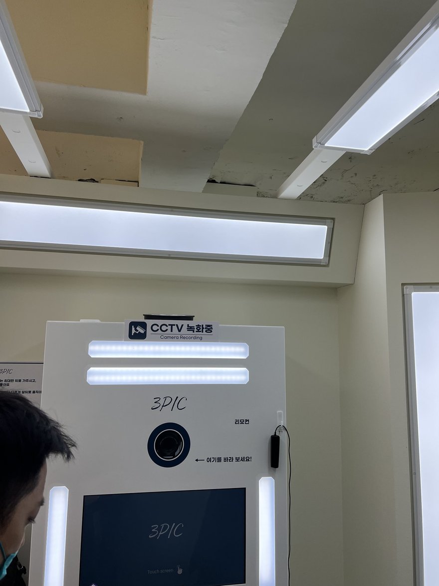 3PIC 店內的白色空間與機台，機台上方貼有 CCTV 녹화중標示，牆面為淺色調