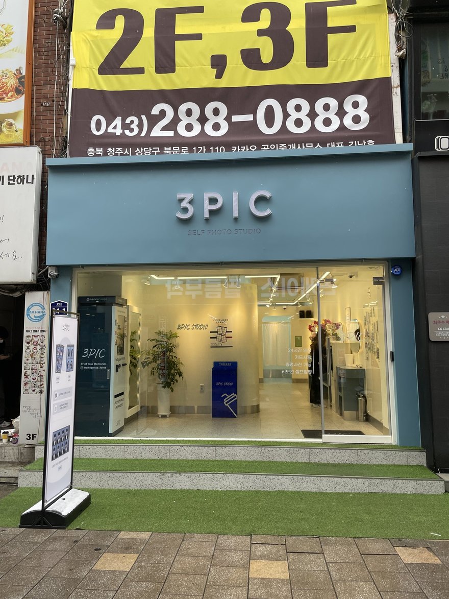 3PIC 清州門市的店面外觀，灰藍色招牌以立體字寫 3PIC 與 SELF PHOTO STUDIO，門前鋪人工草皮，店招上方掛有黃底招租廣告