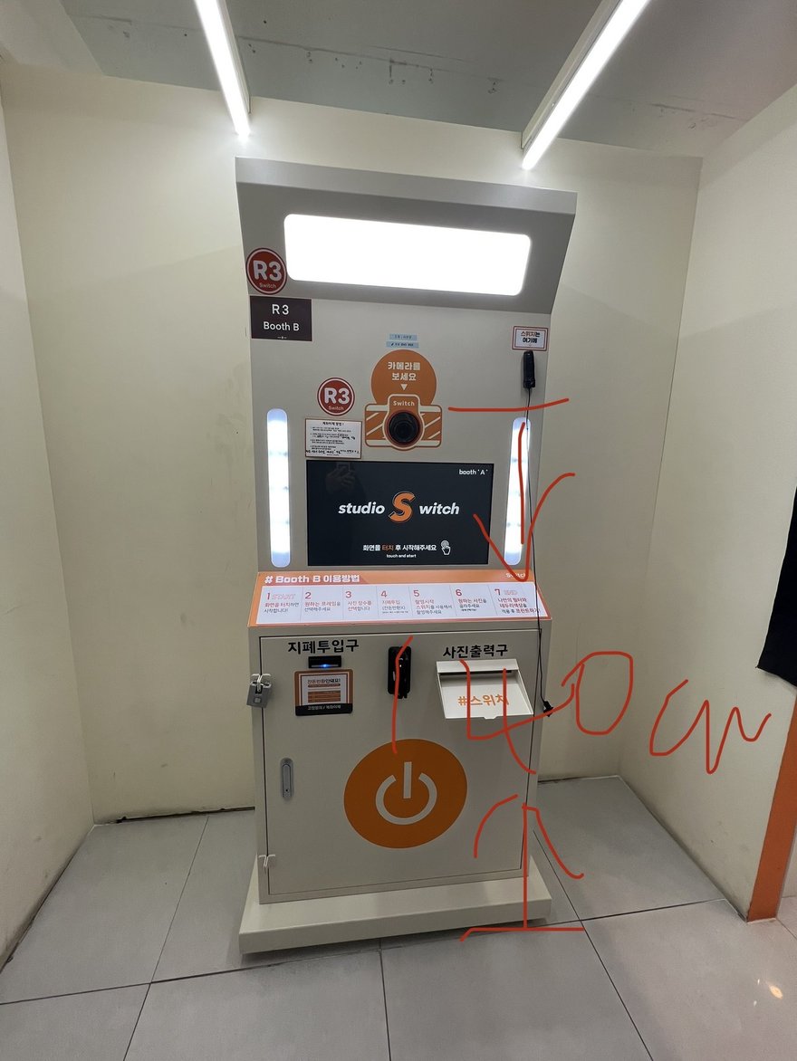 韓式自助拍照機的正面照片，白色機身、面板設橘色電源鍵，照片上留有以紅線畫出的分析標記與箭頭