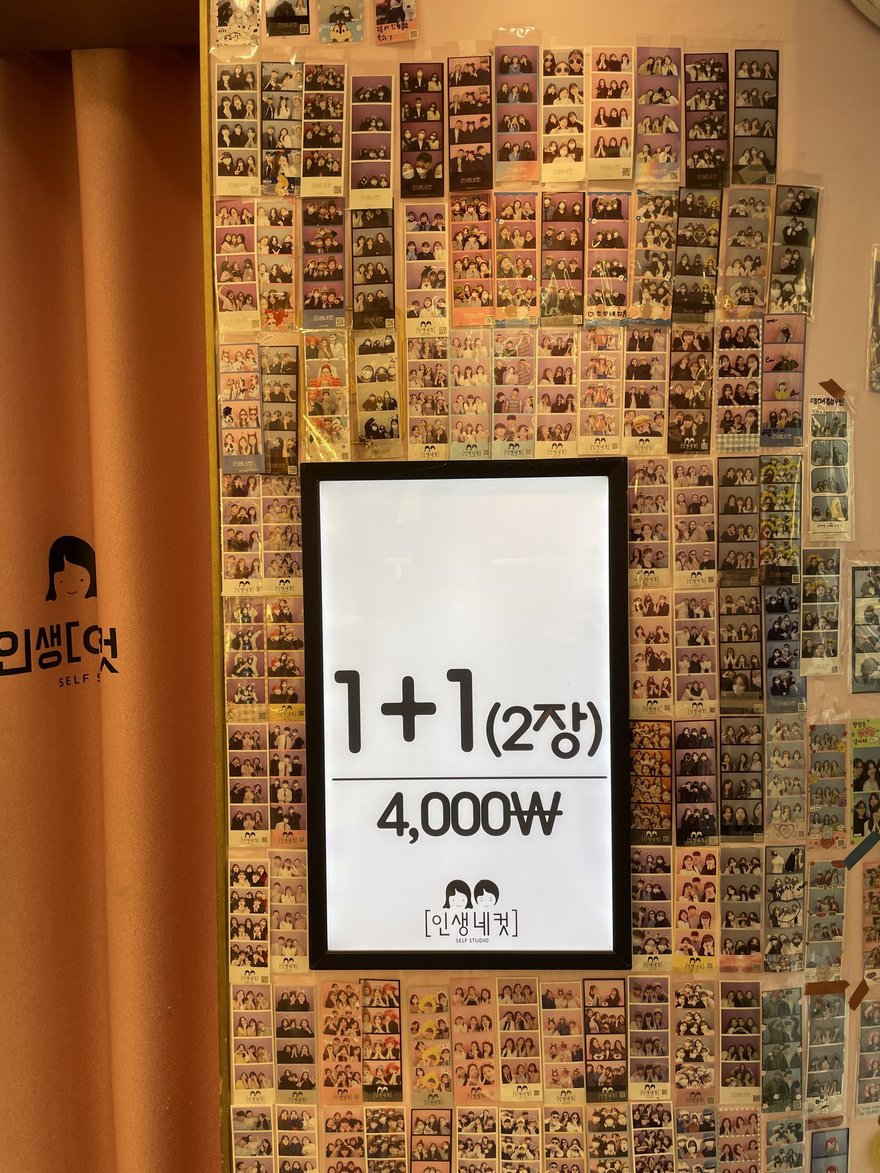 인생네컷店內的價目牌，白底黑字寫 1+1（2장）4,000W，掛在橘色柱體與樣張牆之間