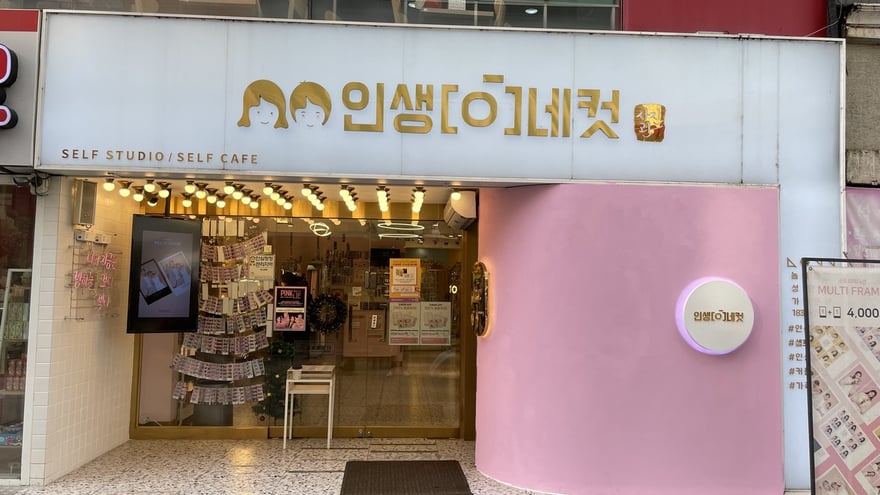 人生四格門市正面，白色招牌寫인생네컷與 SELF STUDIO／SELF CAFE，入口一側是粉紅色弧形牆，門口立著價目看板，店內牆面貼滿樣張。