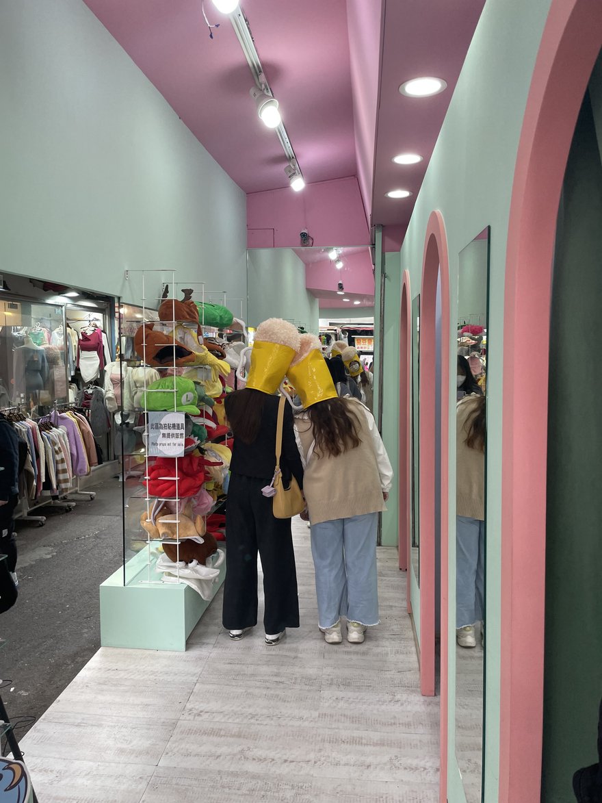台中一中商圈店的道具走道，兩名顧客背對鏡頭、頭上戴著啤酒杯造型頭套，左側層架滿是動物與角色造型頭套並貼有「此區為拍貼機道具 無提供販售 Photo props not for sale」告示，右側粉紅拱門與落地鏡之外可直接看到街面攤商的服飾架與臭豆腐招牌