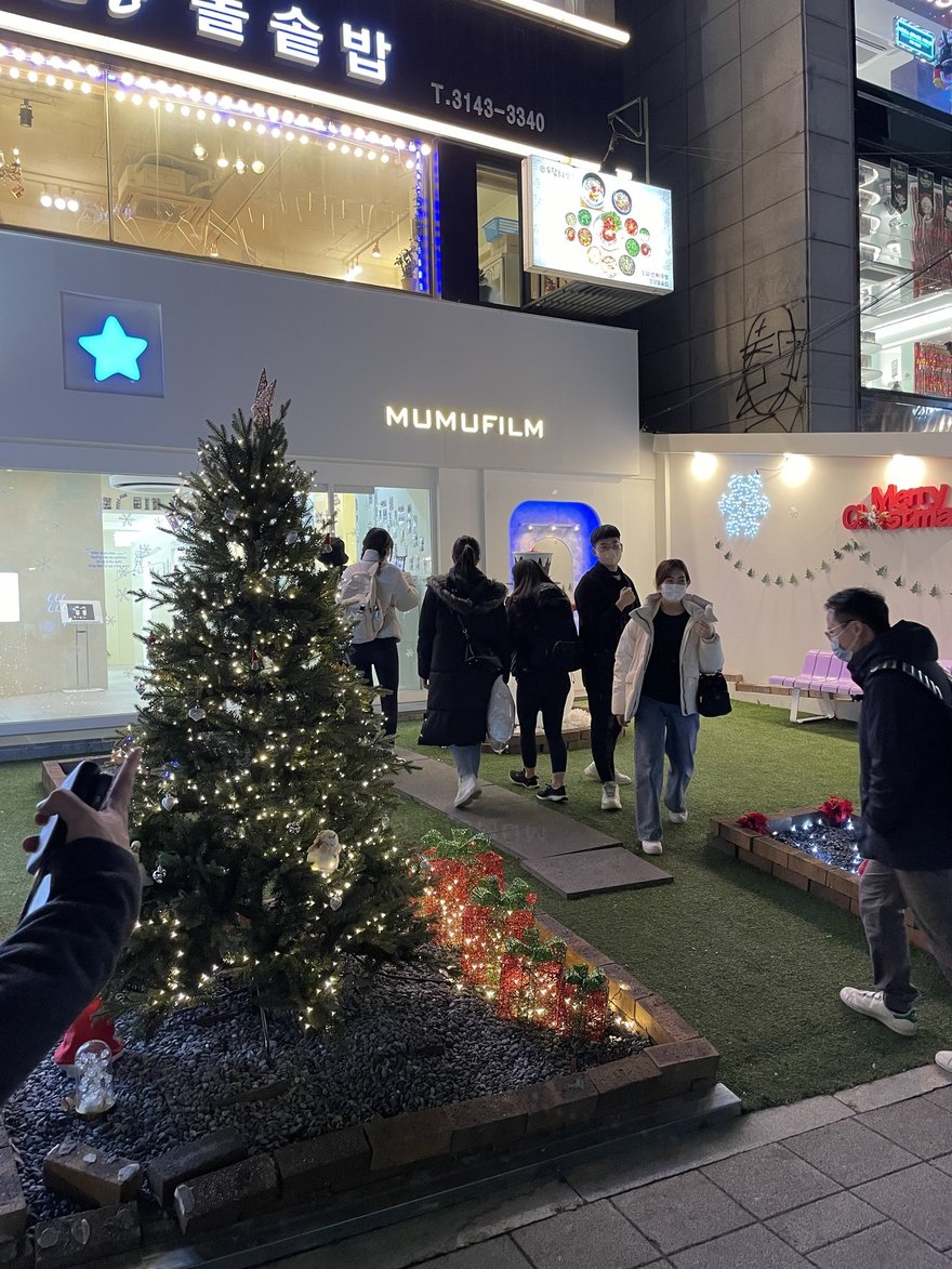 MUMUFILM 的夜間門市外觀，店前為一整片人工草皮前庭，中央立著掛滿燈飾與紅色裝飾球的大型聖誕樹，招牌為藍底白字 MUMUFILM
