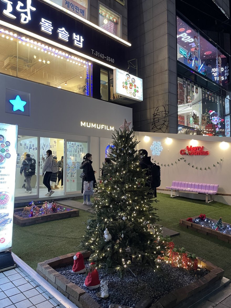 夜晚的商圈街景，MUMUFILM 門市前鋪著人工草皮、立著掛滿燈飾的聖誕樹，兩側是餐廳與其他店家的招牌，行人從店前走過。