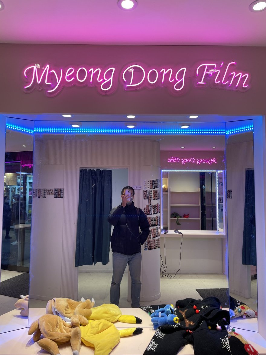 Myeong Dong Film 店內的粉紫色空間，霓虹招牌下方為鏡面與櫃檯，地上堆著成堆的絨毛玩偶道具