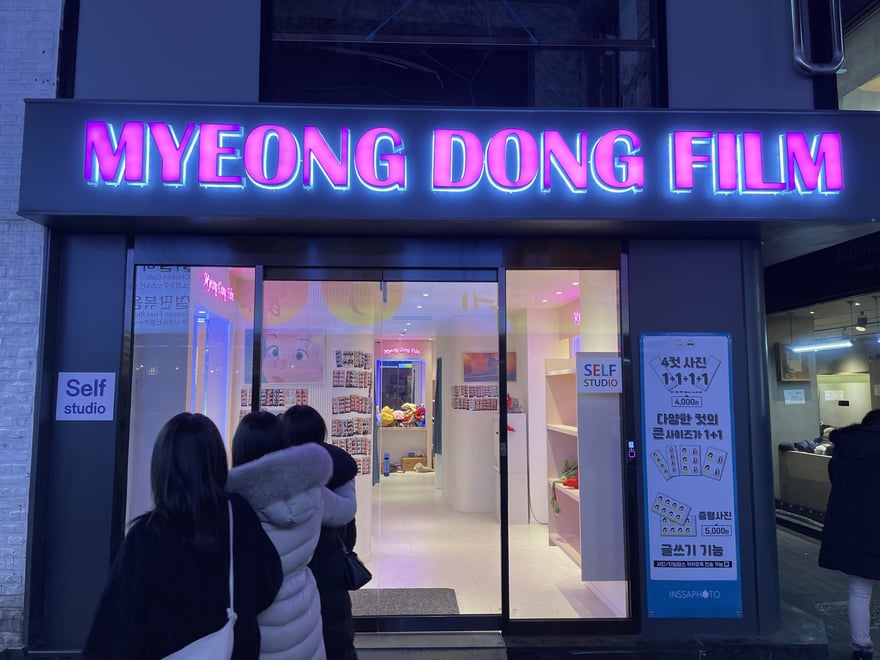 Myeong Dong Film 的夜間店面外觀，粉紫色霓虹招牌寫 MYEONG DONG FILM，兩側標示 Self studio，右側立著直式價目看板，門口有顧客排隊