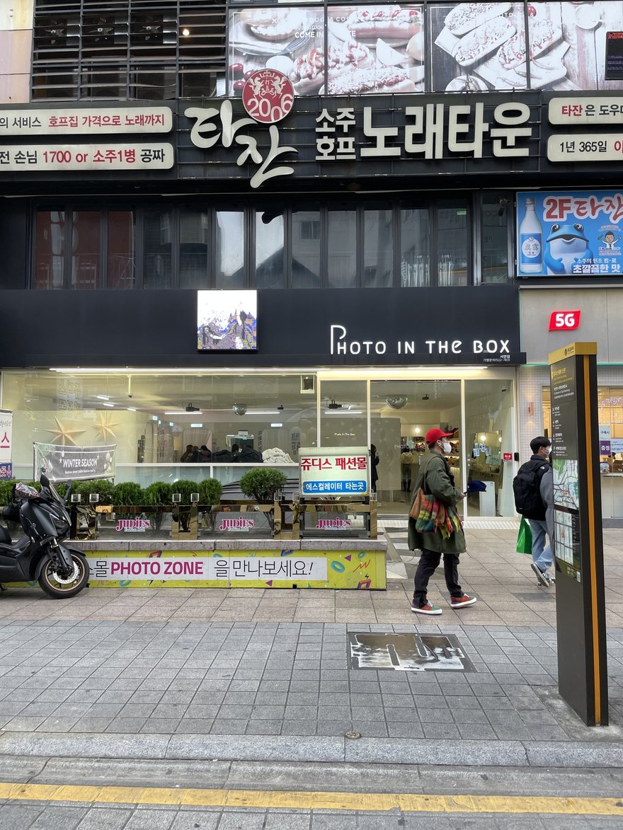 Photo in the Box 釜山西面店的店面外觀，黑色招牌以白色字寫 PHOTO IN THE BOX，右下角印서면점與가맹문의 1522-7831，店面位於소주호프 노래타운樓下，旁邊是쥬디스 패션몰的手扶梯入口指示牌