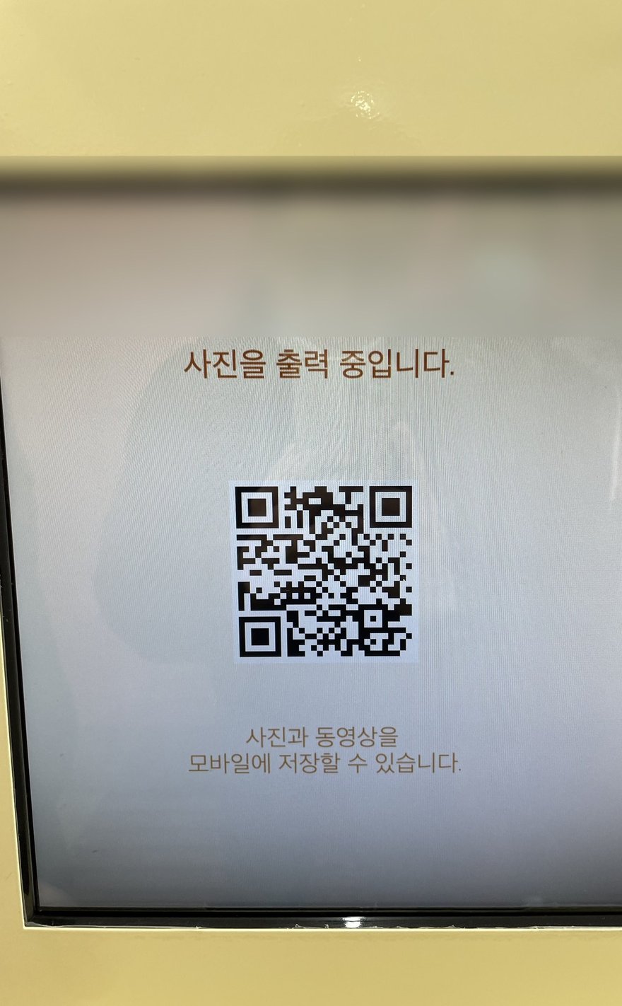 Photo in the Box 的出片等待畫面，螢幕顯示사진을 출력 중입니다照片列印中，中央為 QR code，下方寫사진과 동영상을 모바일에 저장할 수 있습니다，畫面已裁切去識別化