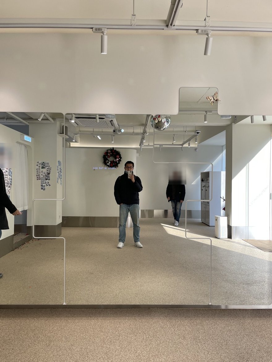Photo in the Box 商場店的入口，挑高白色空間、天花板懸掛鏡球，兩側為商場走道，畫面中路人臉部已遮蔽