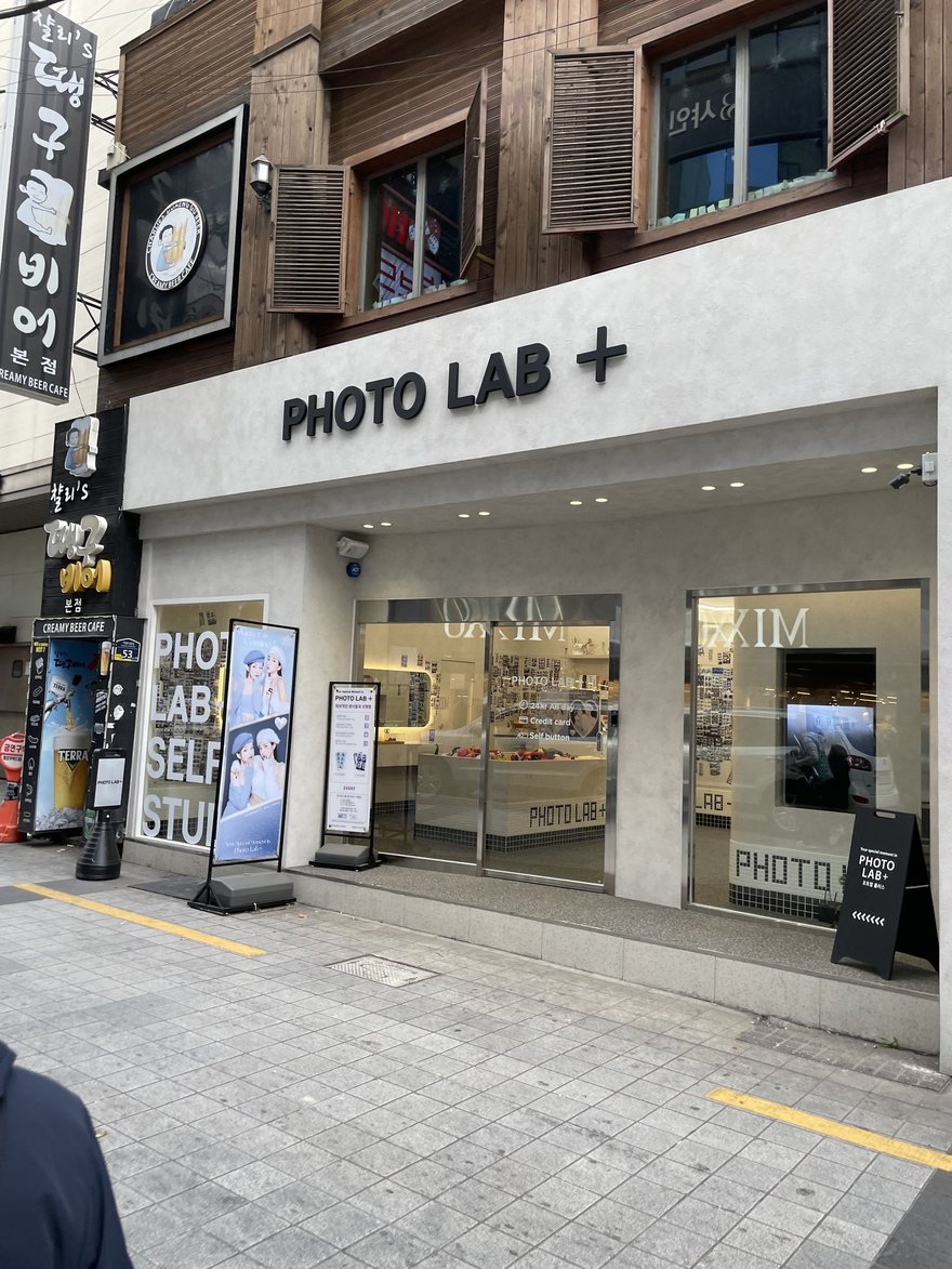 PHOTO LAB+ 的門市外觀，黑色招牌寫 PHOTO LAB +，整面落地玻璃上貼有超大字體的 PHOTO LAB + SELFIE STUDIO