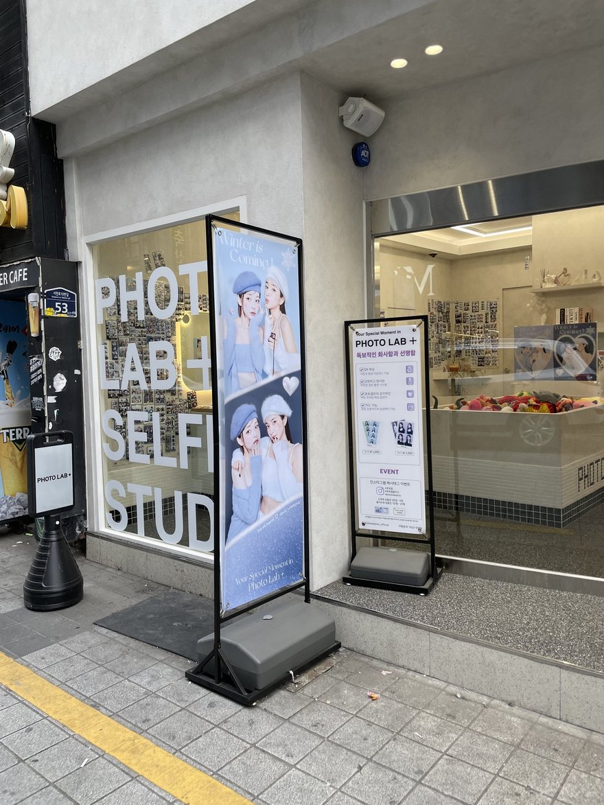 PHOTO LAB+ 門口的直式燈箱立牌與店面入口，燈箱上為模特兒樣張