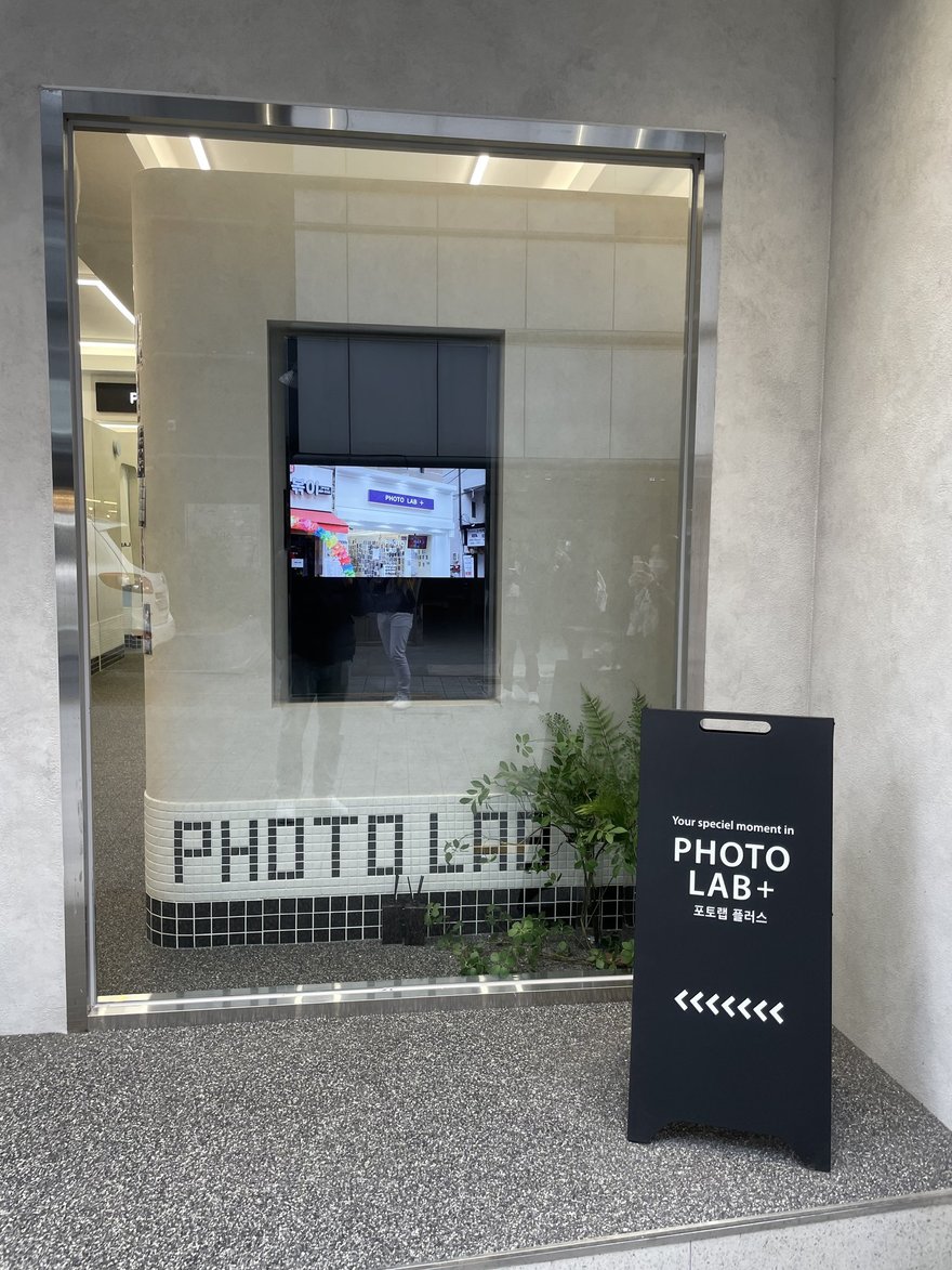 PHOTO LAB+ 店內的入口與機台，玻璃門上印 PHOTO LAB 字樣，門旁立著指示牌