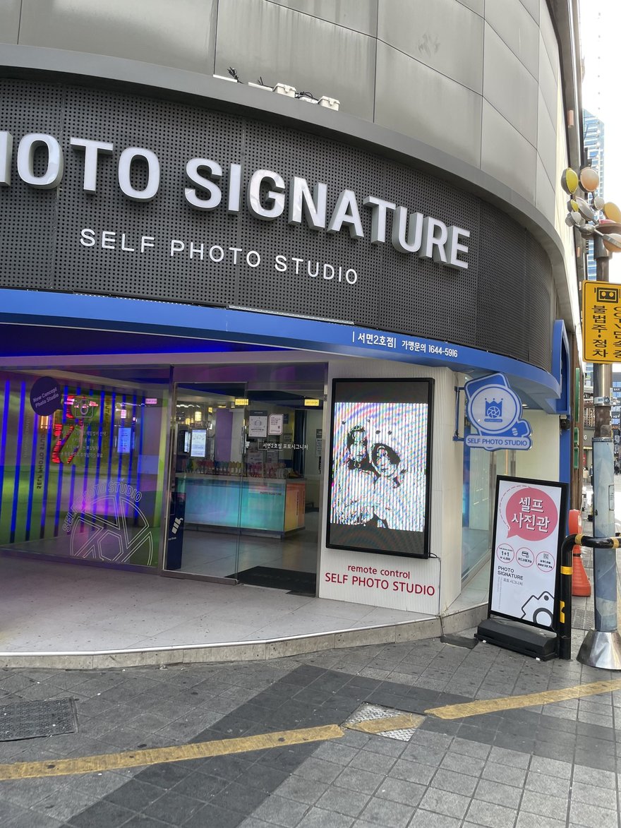 Photo Signature 的街邊門市外觀，黑色招牌以白字寫 PHOTO SIGNATURE 與 SELF PHOTO STUDIO，門口兩側為粉紫色燈光與模特兒燈箱