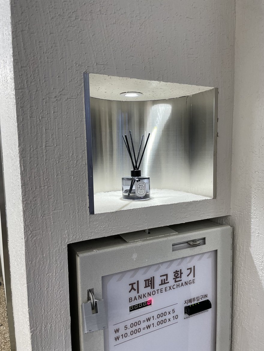 Photo Slate 店內的紙鈔兌換機，面板寫지폐교환기 BANKNOTE EXCHANGE，標示 5,000 韓元換 1,000 韓元五張、10,000 韓元換十張，上方壁龕擺放香氛擴香瓶