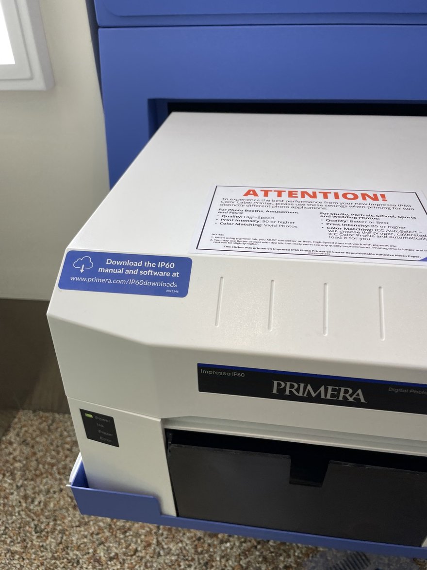 自助拍照機機櫃內的白色 PRIMERA 相片印表機，機頂貼著紅字 ATTENTION 的警告標籤。