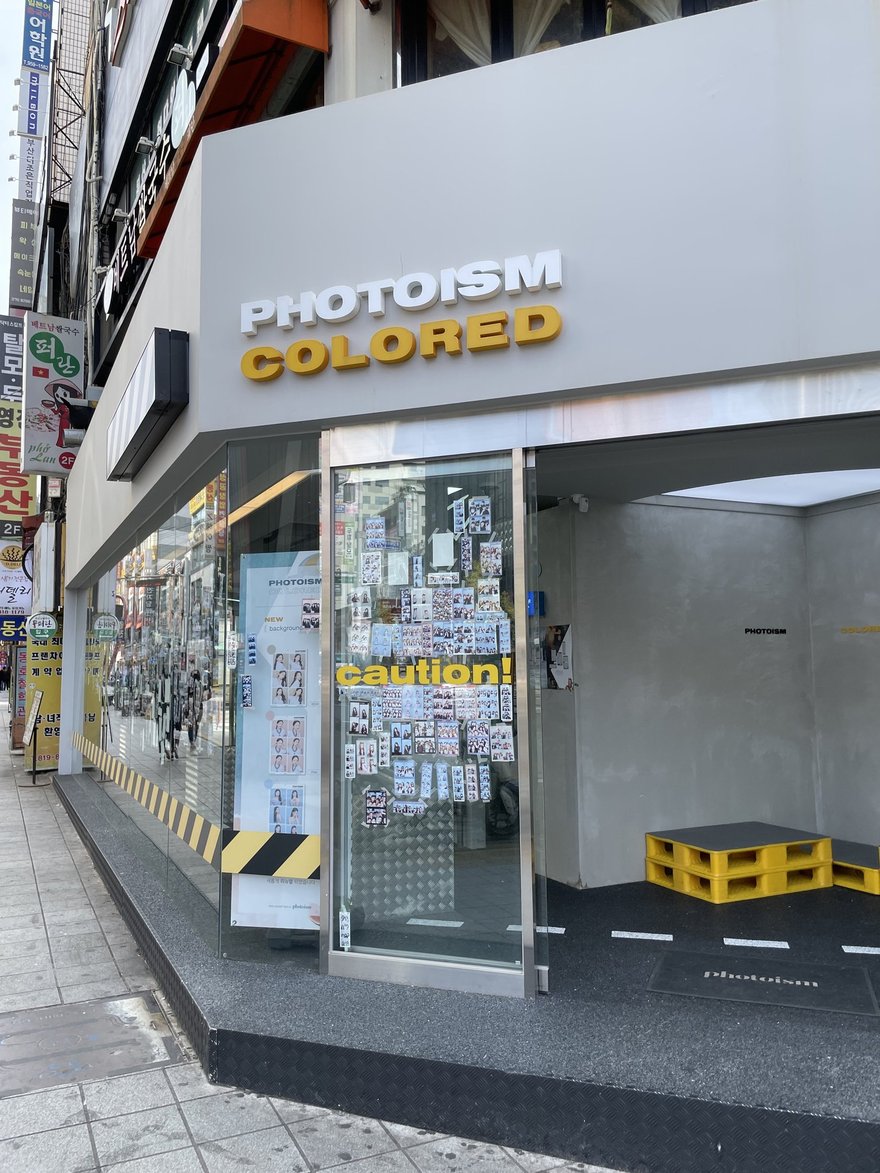 PHOTOISM COLORED 的門市外觀，白色招牌上方寫 PHOTOISM、下方黃字寫 COLORED，玻璃門兩側為黃黑警示條紋與 caution 字樣