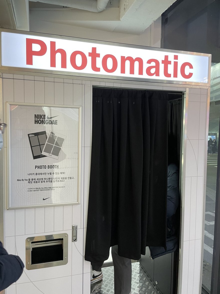 NIKE 弘大店內的 Photomatic 黑白拍照亭，白色亭體上方是紅字 Photomatic 招牌，左側貼著 NIKE HONGDAE 的使用說明與樣張，右側為黑色布簾入口。