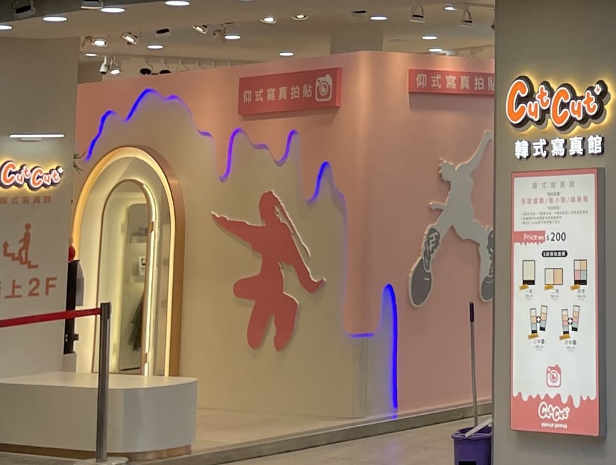 Cut Cut+ 一樓牆面掛著「仰式寫真拍貼」紅色燈箱，牆上是由下往上看、看得見鞋底的人形剪影，左側立牌寫「請上 2F」，右側價目燈箱標示 Price 單場 200 元與五款版型。