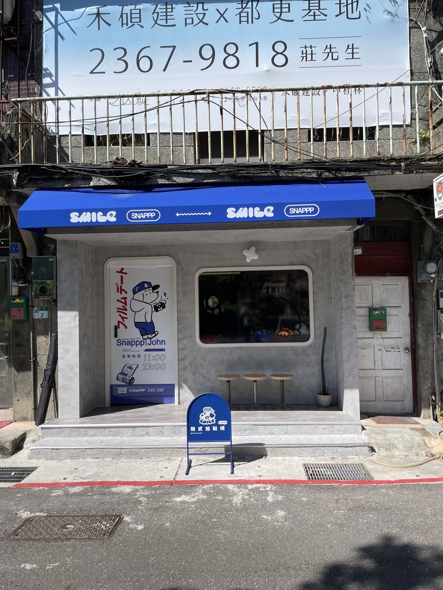 SNAPPP 與 SMILE 聯名的赤峰店店面外觀，藍色雨庇上交錯印著 SMILE 與 SNAPPP 兩個品牌標記，水泥粉光立面配圓角開窗與三張矮凳