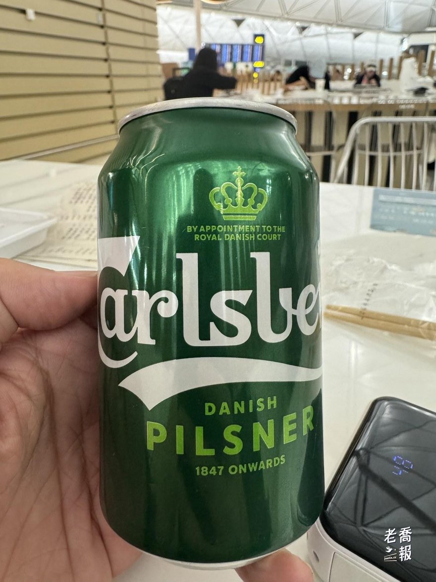 手上拿著一罐 330 毫升的 Carlsberg 啤酒，背景是機場餐廳座位