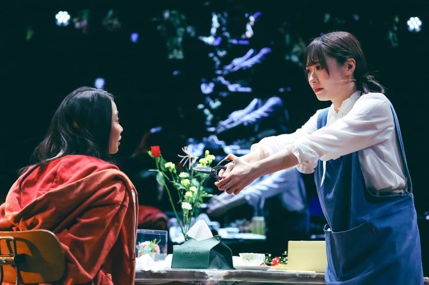 日本版開幕日舞台照，左為飾演妹妹蒼的屋比久知奈，穿藍色圍裙站在花店工作桌前，右為飾演姊姊紅的唯月ふうか，披著紅色外套坐在桌邊