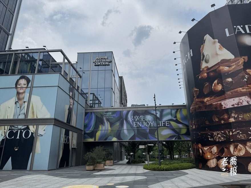 西岸中環 WESTBUND CENTRAL 的玻璃帷幕招牌，右側是包覆整棟轉角的 LÄDERACH 巧克力大型廣告，天橋上掛著「盡享生活 ENJOY LIFE」橫幅，地面廣場幾乎沒有行人
