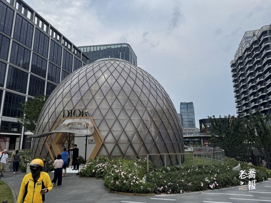 西岸中環二期廣場上的網格球形穹頂，門楣掛著 DIOR 字樣，周圍是整圈花圃，一名穿黃色外送制服的騎手正走過
