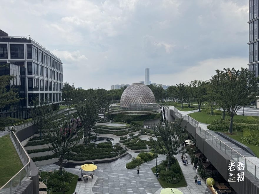 從高處俯瞰西岸中環二期的梯田景觀，層層下降的草坡與步道之間有黃色遮陽傘，中央是網格狀的球形穹頂建築，兩側是新落成的辦公樓