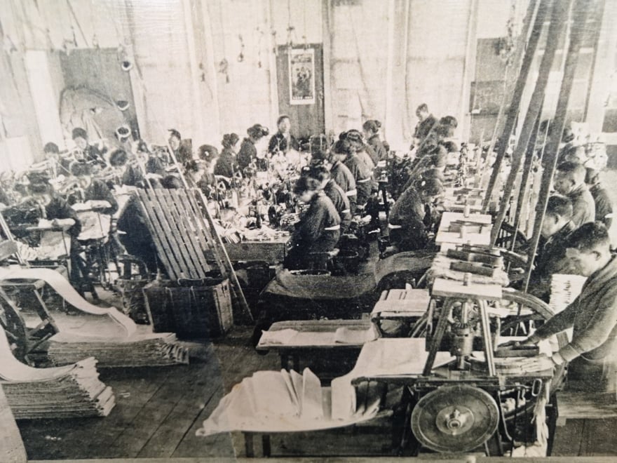 大正時代埼玉縣行田市的足袋裁斷工場黑白照片：長條工作檯兩側坐滿踩著縫紉機的女工，地面與檯面堆疊著裁好的白色足袋布料，右側可見帶皮帶輪的裁斷機具。