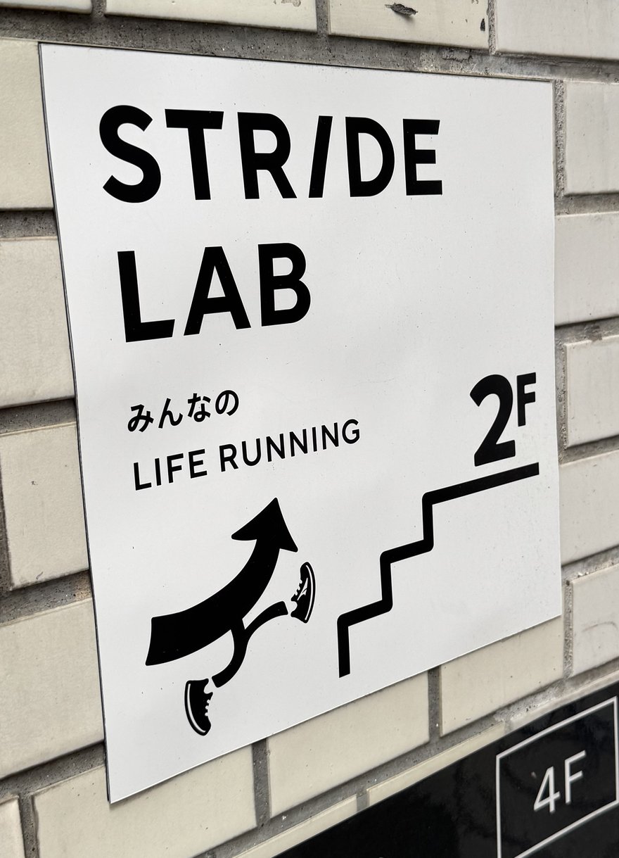 橫濱 STRIDE LAB 店門口的白色指示牌，牌上寫著 STRIDE LAB 與みんなの LIFE RUNNING，右下角以 2F 字樣與一個人跑上樓梯的圖示指引上樓，牌子貼在磚砌外牆上。