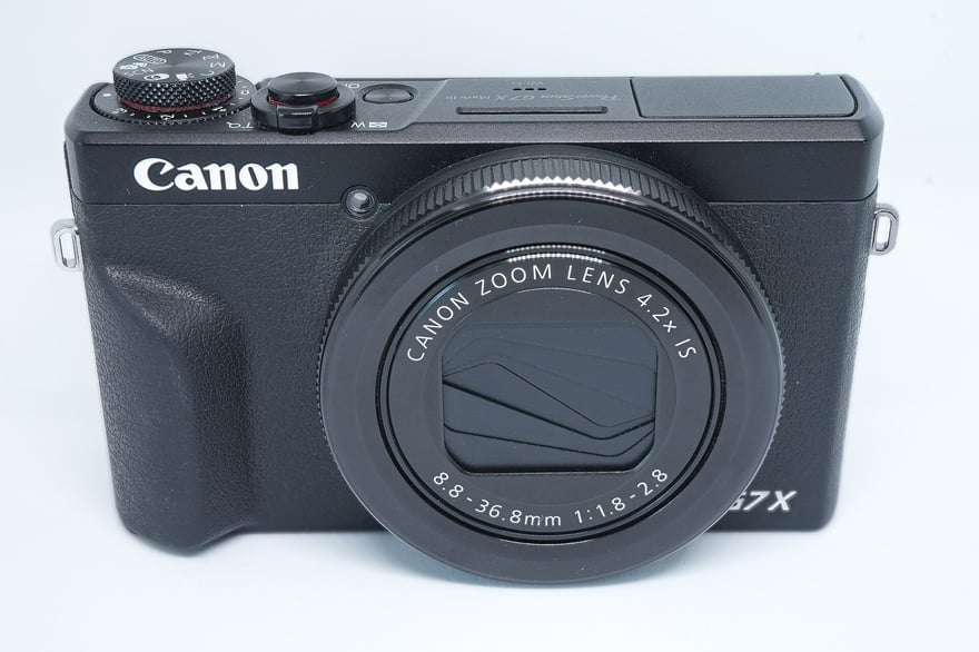 Canon PowerShot G7 X Mark III डिजिटल कॉम्पैक्ट कैमरे का सामने का हिस्सा, काली बॉडी, लेंस रिंग पर CANON ZOOM LENS 4.2x IS और 8.8–36.8mm 1:1.8–2.8 लिखा है; 2026 में ली गई फ़ोटो।