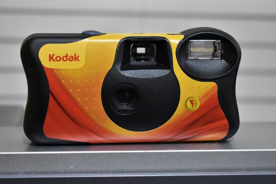 फ़्लैश वाला Kodak डिस्पोज़ेबल कैमरा, सामने से; पीले, नारंगी और लाल काग़ज़ी कवर पर Kodak का लोगो और बिजली का निशान, बीच में फ़िक्स्ड फ़ोकस वाला छोटा लेंस, ऊपर बाईं ओर व्यूफ़ाइंडर और ऊपर दाईं ओर फ़्लैश; 2018 में ली गई फ़ोटो।