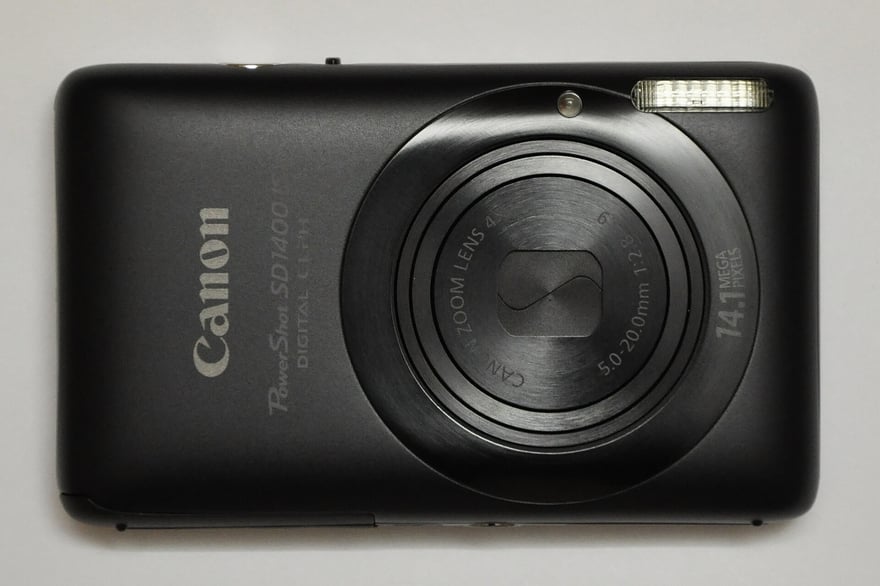Canon PowerShot SD1400 IS का सामने से फ़ोटो: काली, पतली बॉडी वाला पॉकेट डिजिटल कैमरा, लेंस कवर बंद, बॉडी के दाईं ओर 14.1 Mega Pixels लिखा है, बाईं ओर Canon और PowerShot SD1400 IS।