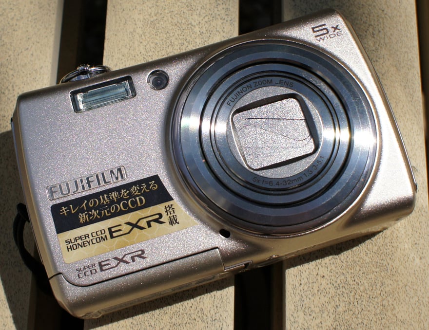 Fujifilm FinePix F200EXR का तिरछा फ़ोटो: शैम्पेन-गोल्ड रंग की बॉडी वाला पॉकेट डिजिटल कैमरा, ऊपर दाएँ 5x WIDE लिखा है, बॉडी के सामने जापानी लेबल लगे हैं जिनका आशय है “सुंदरता का पैमाना बदलने वाला नए आयाम का CCD” और “SUPER CCD EXR से लैस”, और कैमरा हल्के रंग की लकड़ी की बेंच पर रखा है।