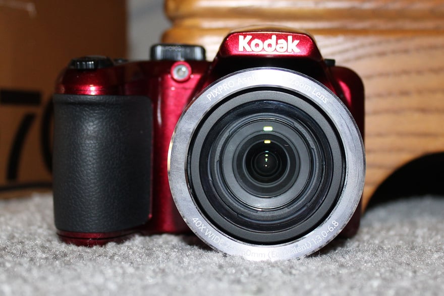 Kodak PixPro AZ401 का सामने से फ़ोटो: लाल बॉडी और काले ग्रिप वाला DSLR जैसी शक्ल का डिजिटल कैमरा, ऊपर सफ़ेद Kodak लिखा है, लेंस रिंग पर 40 गुना वाइड ज़ूम लिखा है, और कैमरा हल्के रंग के कालीन पर रखा है।