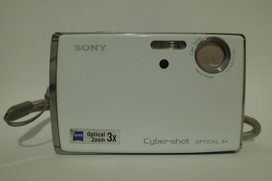 Sony Cyber-shot DSC-T33 का सामने से फ़ोटो: सफ़ेद पतली बॉडी, ऊपर दाएँ कोने में Carl Zeiss Vario-Tessar लेंस, नीचे बाएँ कोने में Zeiss Optical Zoom 3x का लेबल, और बॉडी के नीचे Cyber-shot लिखा है।