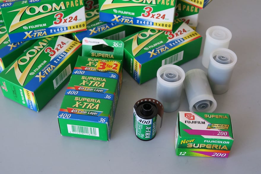मेज़ पर बिखरे हरे पैकेट वाले Fujifilm Superia X-TRA 400 के तीन-रोल और एक-रोल वाले कई डिब्बे, एक Superia 200 का डिब्बा, बगल में कुछ सफ़ेद फ़िल्म कनस्तर और कैसेट से बाहर निकली X-TRA 400 का एक रोल।
