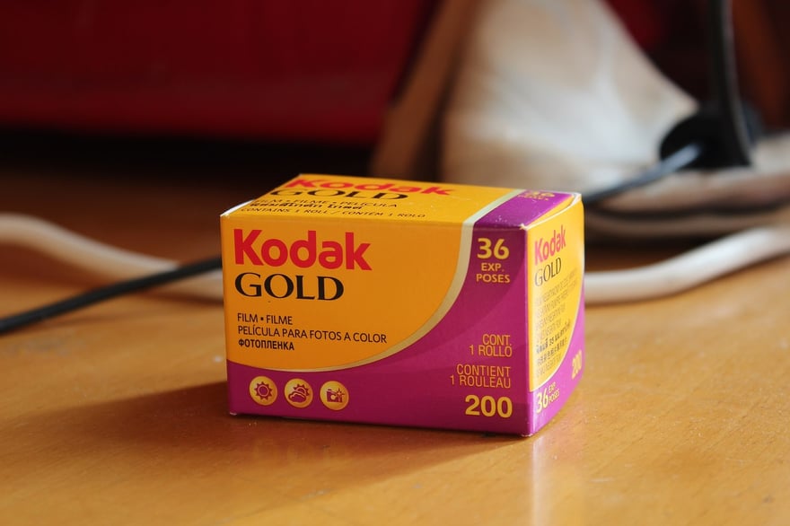 लकड़ी की मेज़ पर खड़ा Kodak Gold 200 कलर नेगेटिव फ़िल्म का एक डिब्बा, पीले और मैजेंटा पैकेट पर Kodak GOLD, 36 एक्सपोज़र और ISO 200 छपा है।