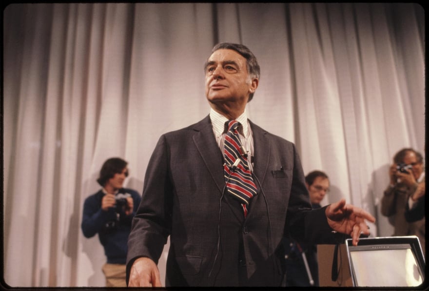Polaroid के संस्थापक Edwin Land लगभग 1971 में सफ़ेद पर्दे के सामने खड़े हैं, गहरे स्लेटी सूट और लाल-नीली धारीदार टाई में, सीने पर माइक्रोफ़ोन लगा है; नीचे दाईं ओर एक Polaroid उपकरण है और पीछे कैमरा लिए दो प्रेस फ़ोटोग्राफ़र हैं।