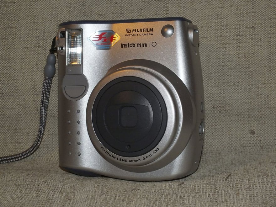 चाँदी के रंग का Fujifilm instax mini 10 सामने से: बड़े गोल लेंस के चारों ओर FUJINON LENS 60mm लिखा है, ऊपर दाईं ओर FUJIFILM INSTANT CAMERA और instax mini 10, ऊपर बाईं ओर फ़्लैश।