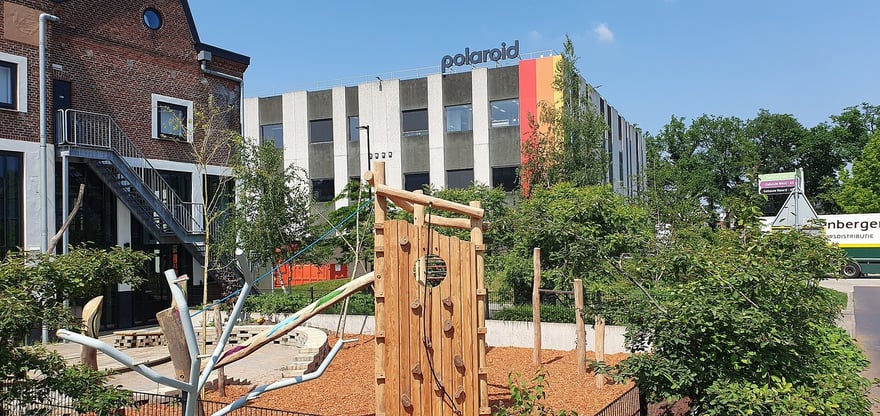 मई 2024 में नीदरलैंड्स के Enschede में Polaroid फ़ैक्ट्री परिसर: स्लेटी-सफ़ेद दफ़्तर की इमारत की छत पर polaroid लिखा है और दीवार पर लाल, नारंगी और पीली रंगीन पट्टियाँ हैं; सामने ईंट की इमारत के पास लकड़ी का बच्चों का झूला है।