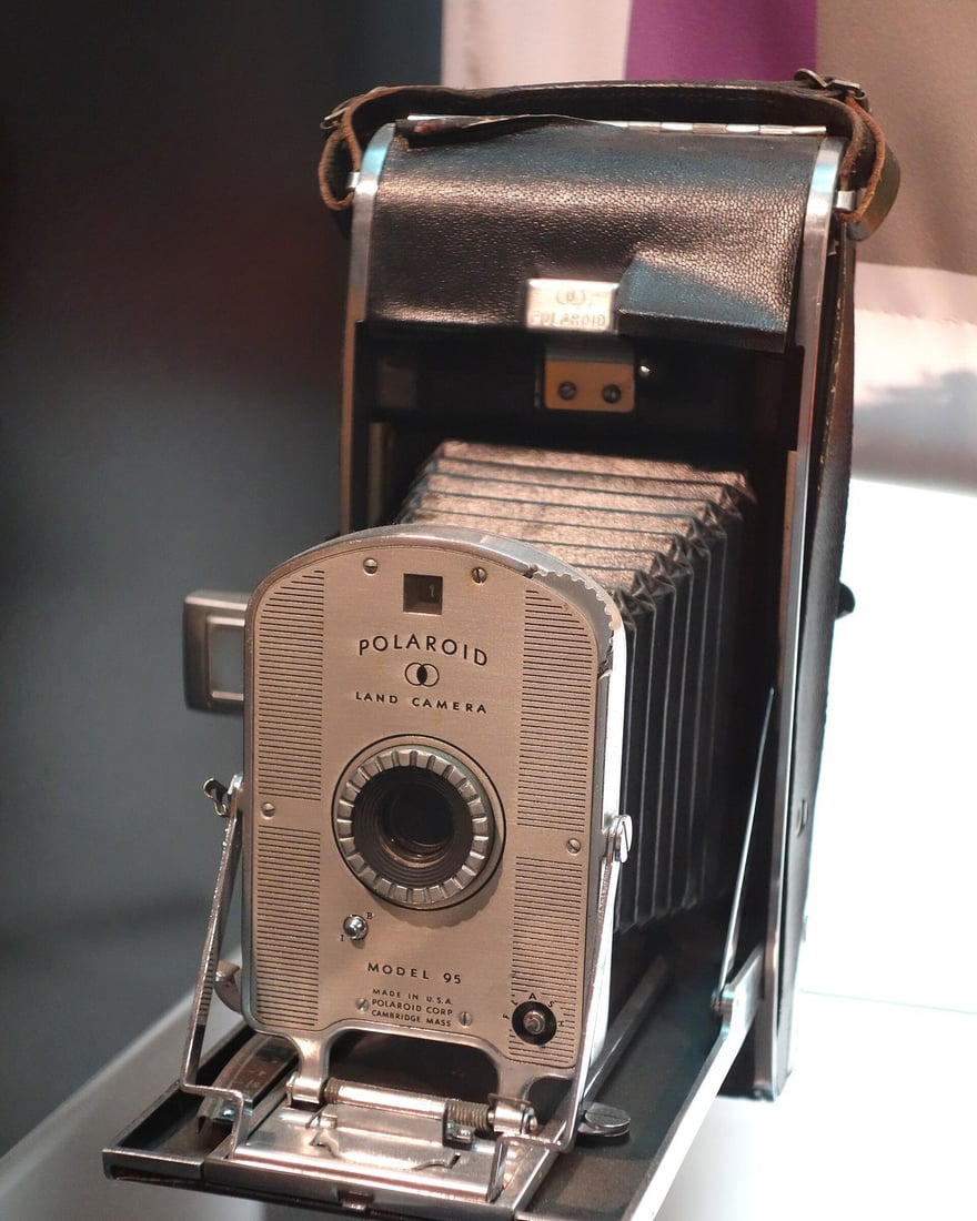 MIT म्यूज़ियम (कैम्ब्रिज, मैसाचुसेट्स) के शोकेस में रखा Polaroid Land Camera Model 95: फ़ोल्ड होने वाली बॉडी से काला धौंकनी जैसा हिस्सा बाहर निकला है, और लेंस प्लेट पर POLAROID LAND CAMERA और MODEL 95 लिखा है।