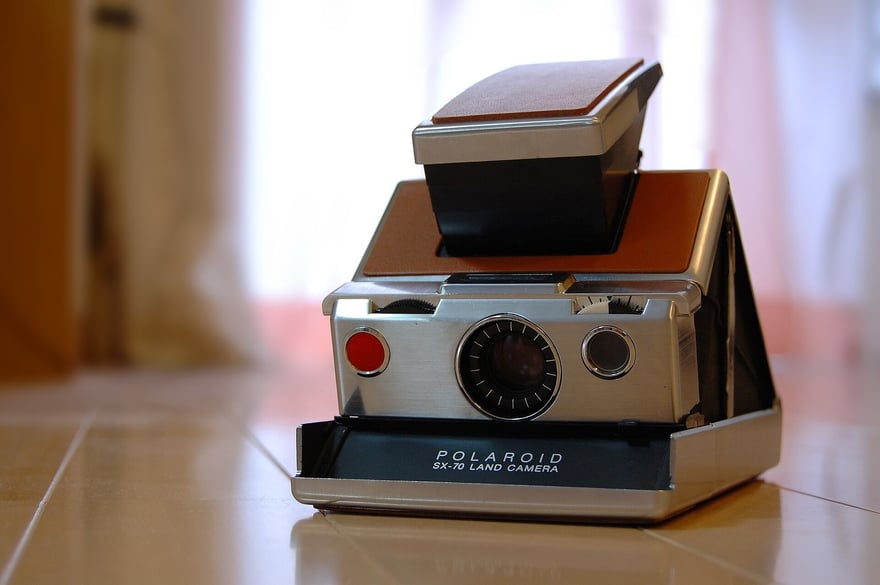 फ़र्श की हल्के रंग की टाइलों पर खुला हुआ खड़ा Polaroid SX-70 Land Camera: ऊपर मुड़ा हुआ व्यूफ़ाइंडर, स्टेनलेस स्टील और भूरे चमड़े की बॉडी, और नीचे फ़ोटो निकलने वाली जगह पर POLAROID SX-70 LAND CAMERA लिखा है।