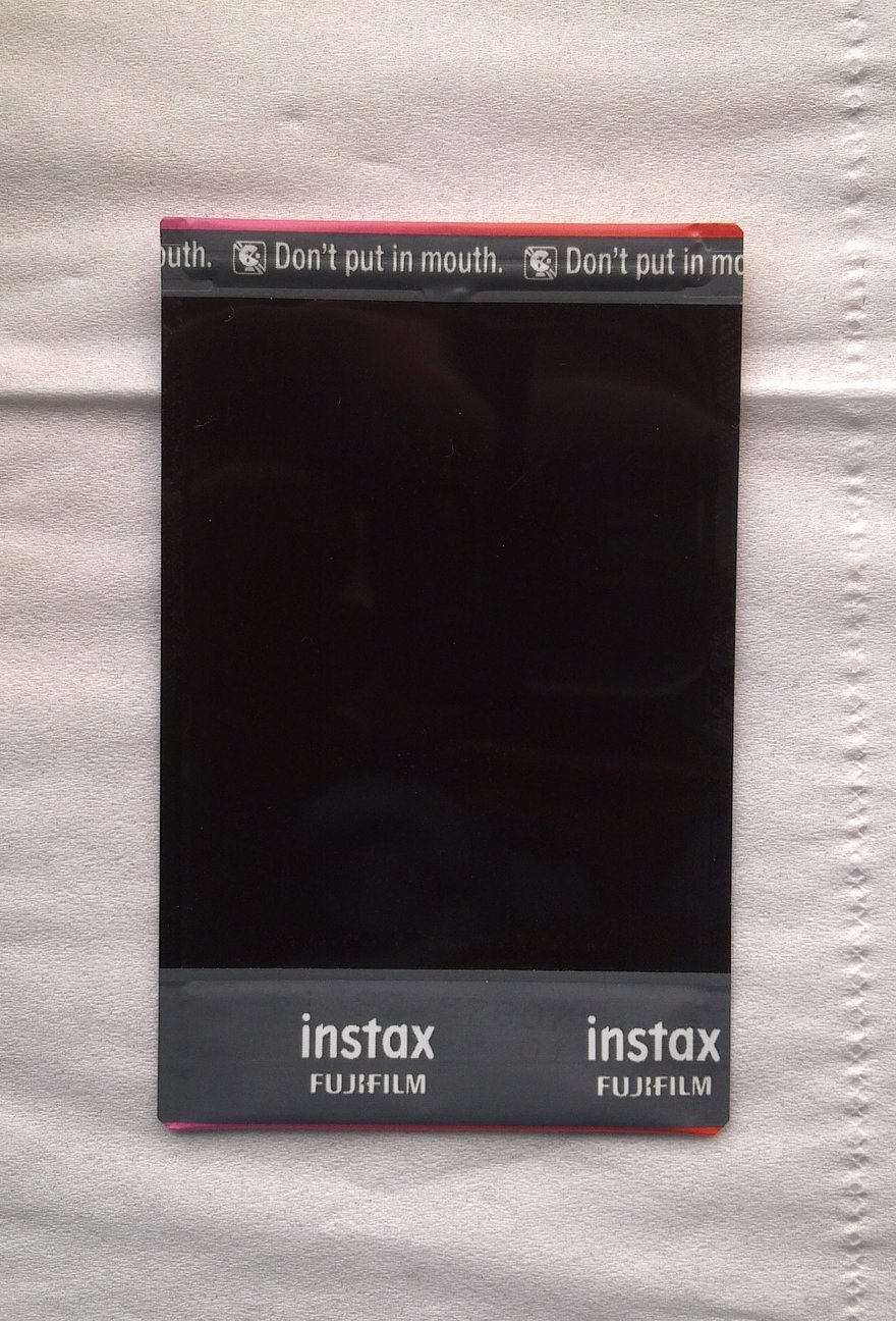 instax mini रेनबो फ़्रेम फ़िल्म का पिछला हिस्सा: काली सतह, ऊपरी किनारे पर बार-बार अंग्रेज़ी चेतावनी “Don’t put in mouth.” छपी है, नीचे instax FUJIFILM लिखा है, और यह सफ़ेद कपड़े पर सपाट रखी है।