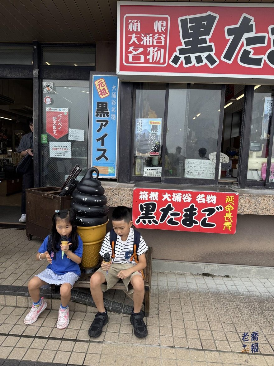 嘻嘻與哈哈坐在極楽茶屋門口的長椅上吃黑色霜淇淋，背後是黑霜淇淋模型與「箱根大涌谷名物 黒たまご」的紅色招牌