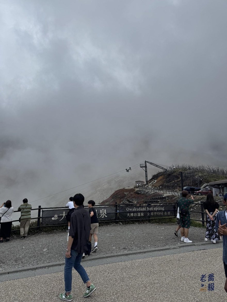 大涌谷觀景台，遊客站在「大涌谷温泉 Owakudani hot spring」木牌前，整片谷地被濃霧籠罩，纜車鋼索伸進霧中