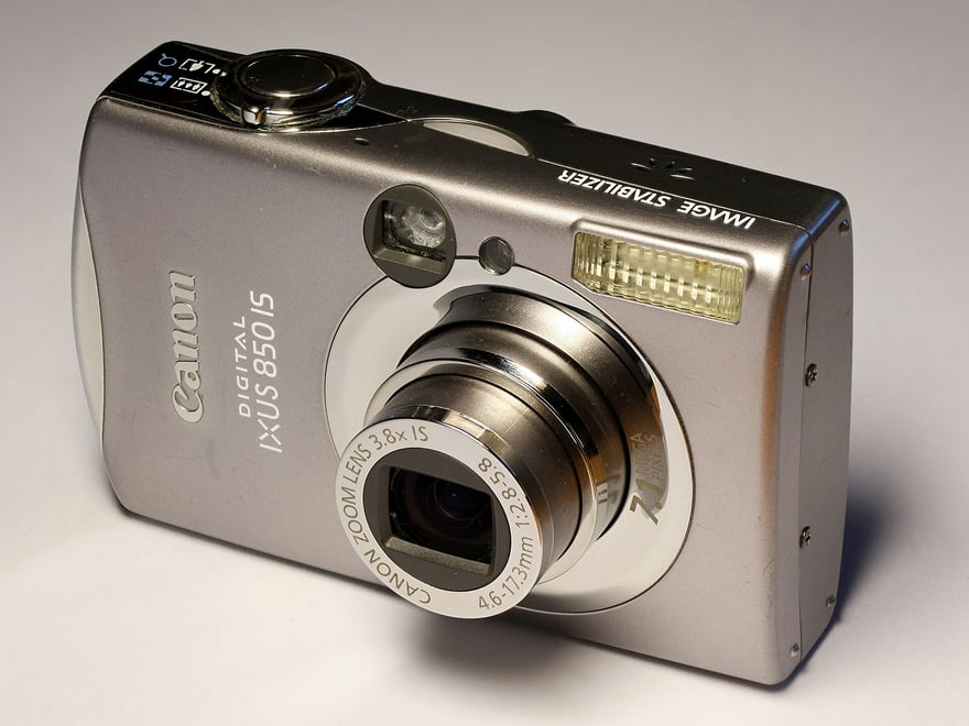 Canon Digital IXUS 850 IS की तिरछी फ़ोटो: चाँदी जैसी धातु की बॉडी वाला जेब में आने लायक़ डिजिटल कैमरा, लेंस रिंग पर Canon Zoom Lens 3.8x IS 4.6–17.3mm लिखा है, ऊपर Image Stabilizer छपा है।
