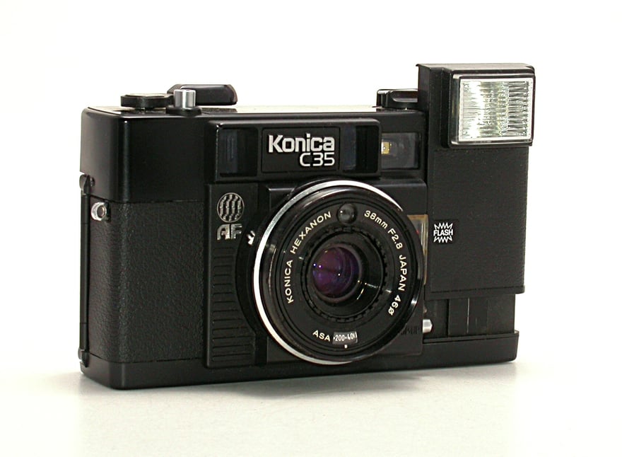 Konica C35 AF की सामने से फ़ोटो: काली बॉडी, लेंस रिंग पर Konica Hexanon 38mm F2.8 खुदा है, बाईं ओर AF का निशान, ऊपर दाईं ओर उठा हुआ बिल्ट-इन फ़्लैश, सफ़ेद बैकग्राउंड।