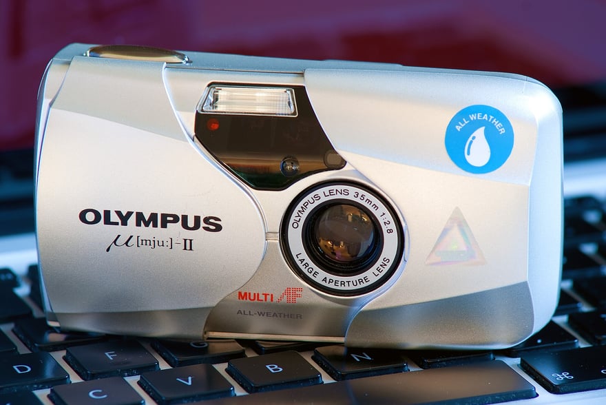 Olympus mju-II की सामने से फ़ोटो: चाँदी जैसी, कैप्सूल आकार की चिकनी बॉडी, स्लाइडिंग कवर खुला है और Olympus Lens 35mm 1:2.8 लिखा लेंस दिख रहा है, ऊपर दाईं ओर नीले रंग का All Weather (पानी की बौछार से सुरक्षा) निशान; कैमरा कीबोर्ड पर रखा है।